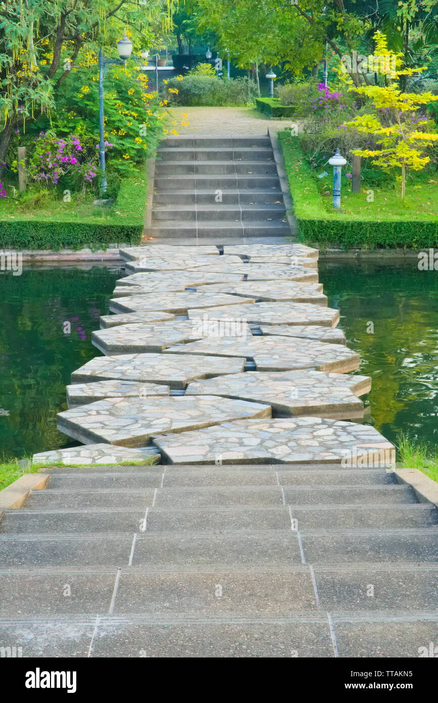 A portrait view of cement steps, leading down towards a green canal stone bridge with watery gaps, and beyond into a lovely, flowery garden park. Stock Photo