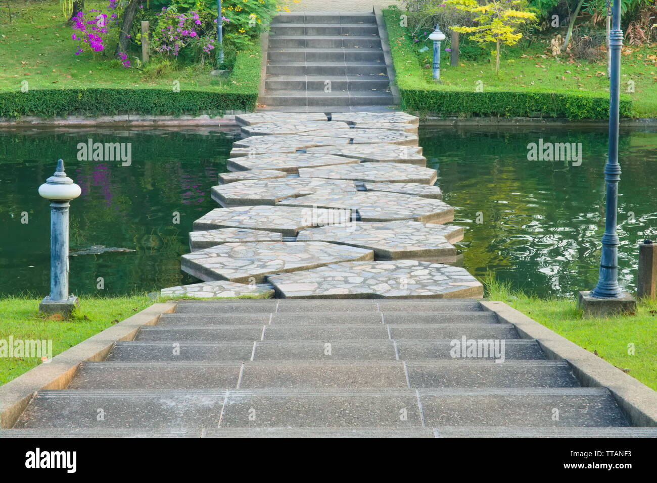 A landscape view of cement steps, leading down towards a green canal stone bridge with watery gaps, and beyond into a lovely, flowery garden park. Stock Photo