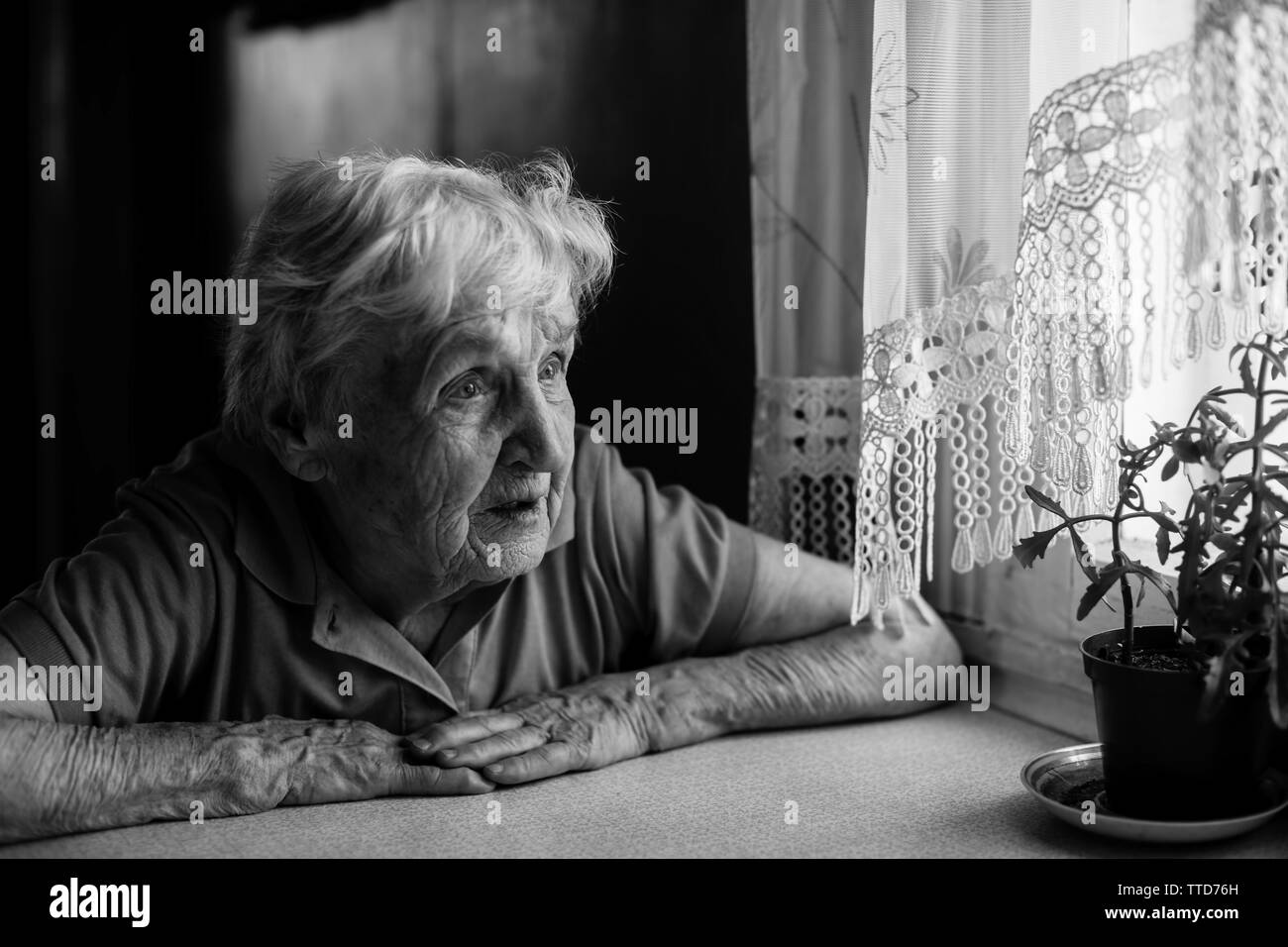 Old lonely woman sitting near the window in his house. Black and white ...