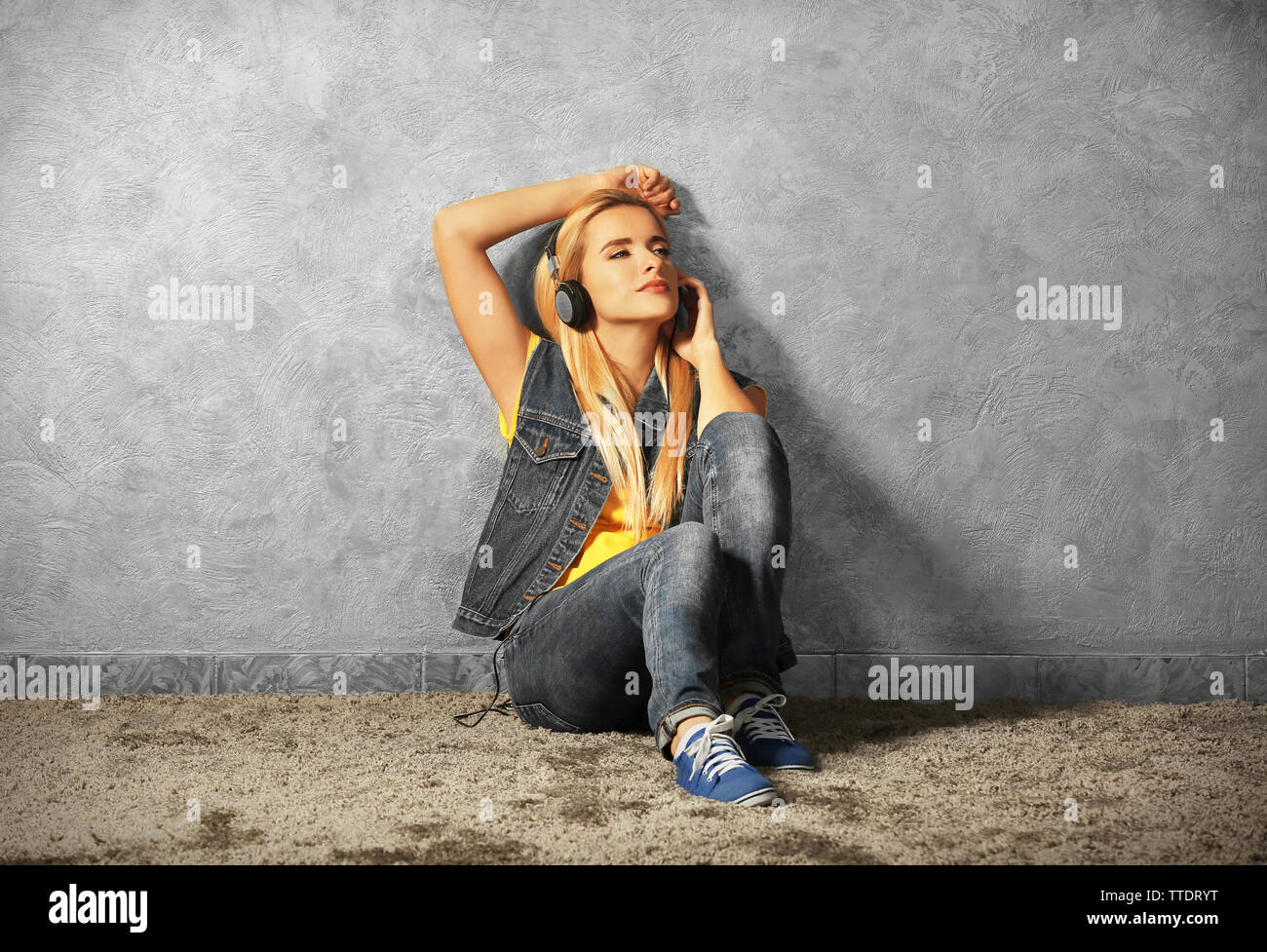Young woman sitting on the floor and listening to music on a grey wall ...