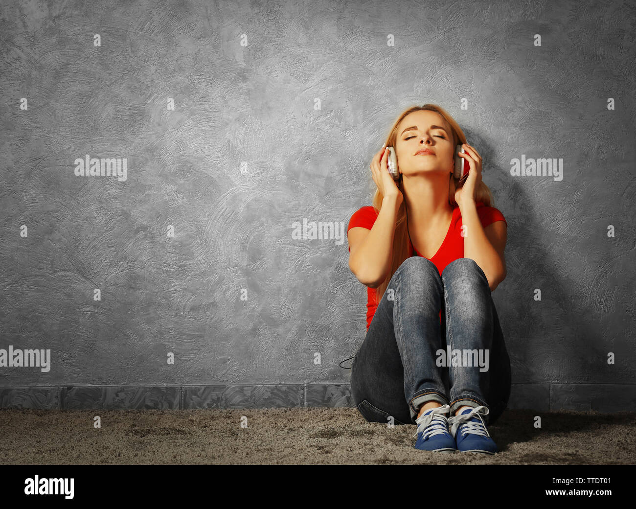Young woman sitting on the floor and listening to music on a grey wall ...