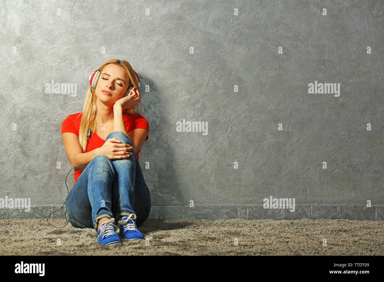 Young woman sitting on the floor and listening to music on a grey wall ...