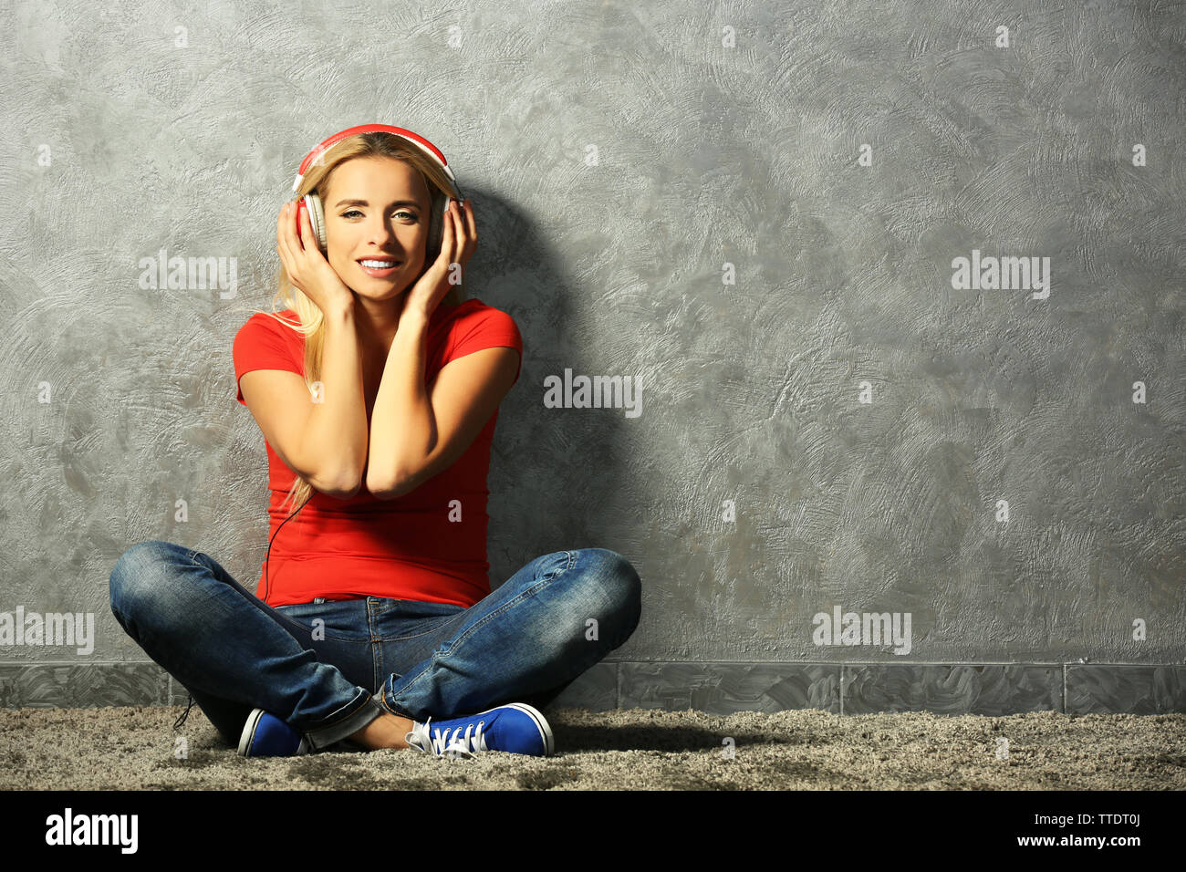 Young woman sitting on the floor and listening to music on a grey wall ...