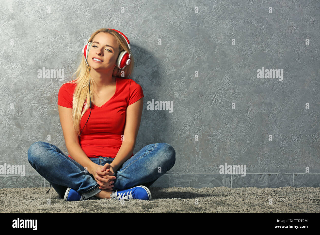 Young woman sitting on the floor and listening to music on a grey wall ...