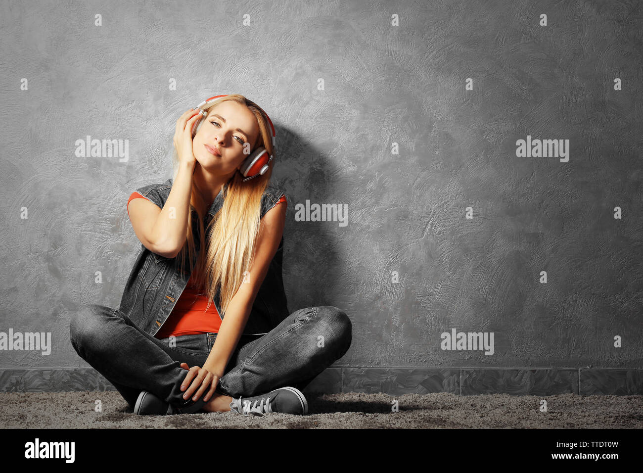 Young woman sitting on the floor and listening to music on a grey wall ...