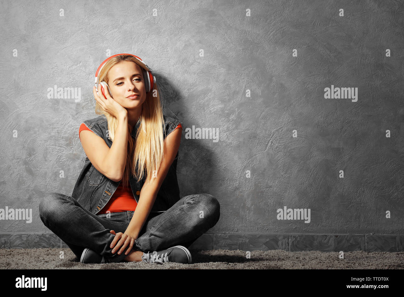 Young woman sitting on the floor and listening to music on a grey wall ...