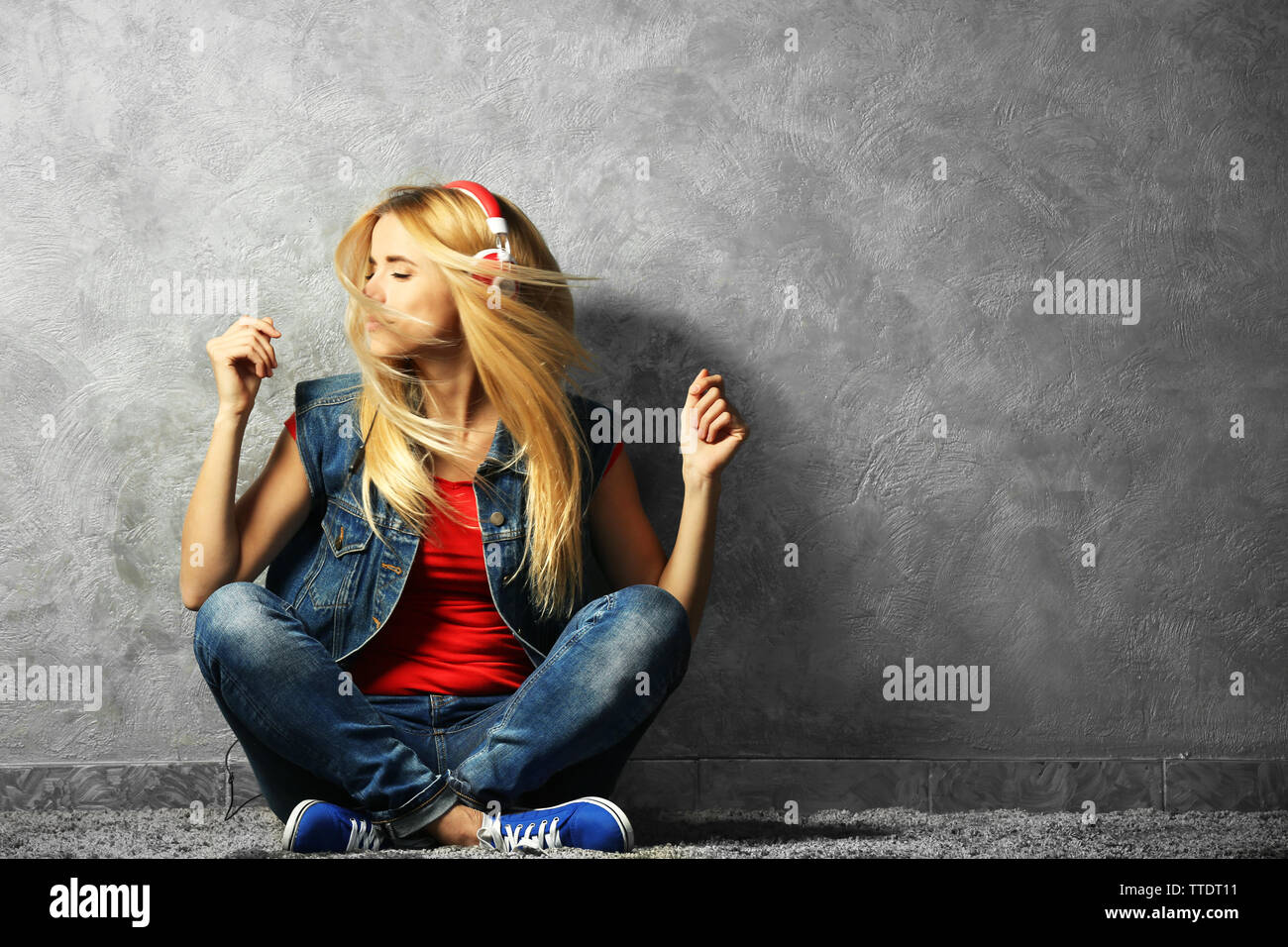 Young woman sitting on the floor and listening to music on a grey wall ...