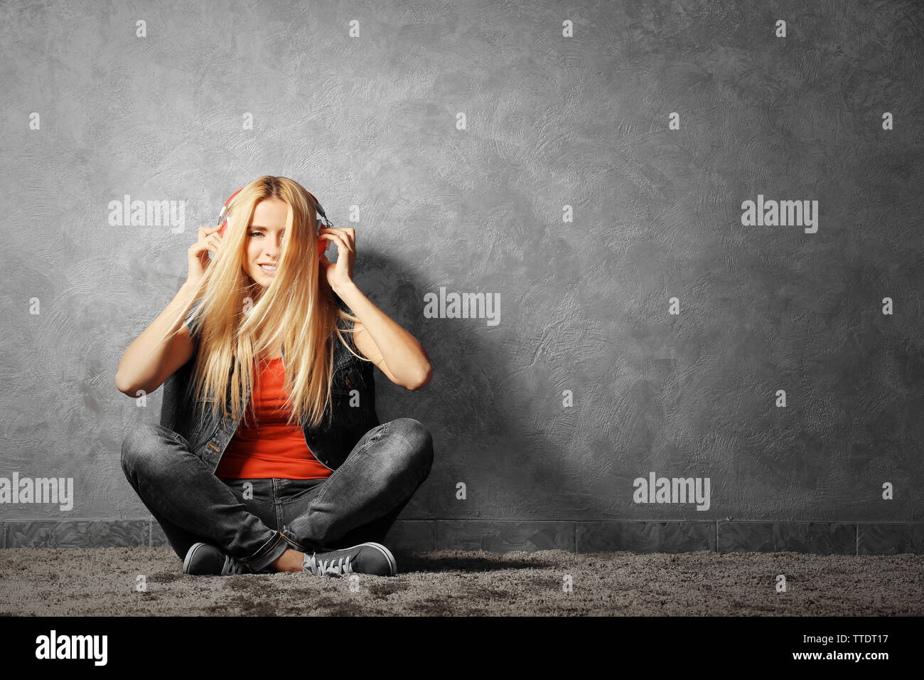 Young woman sitting on the floor and listening to music on a grey wall ...