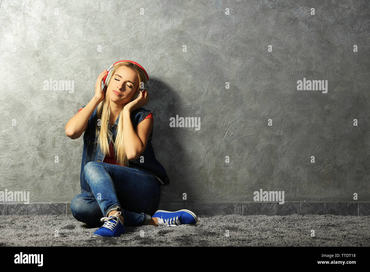 Young woman sitting on the floor and listening to music on a grey wall ...
