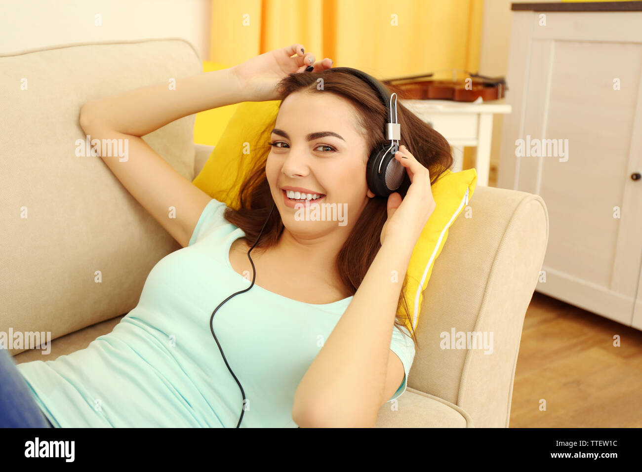 Happy young woman with headphones listening to music on a sofa at home ...