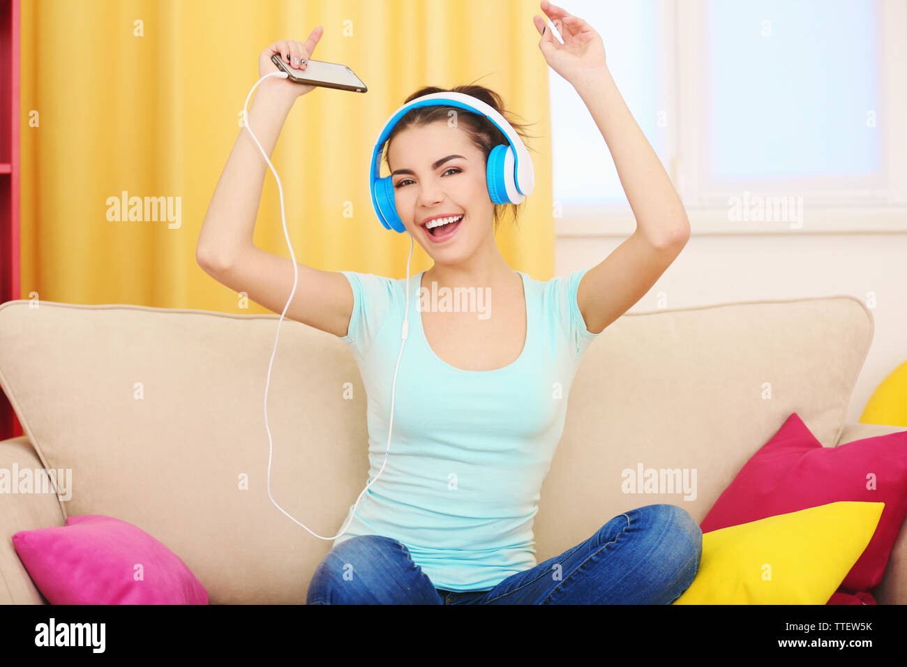 Happy young woman with headphones listening to music on a sofa at home ...