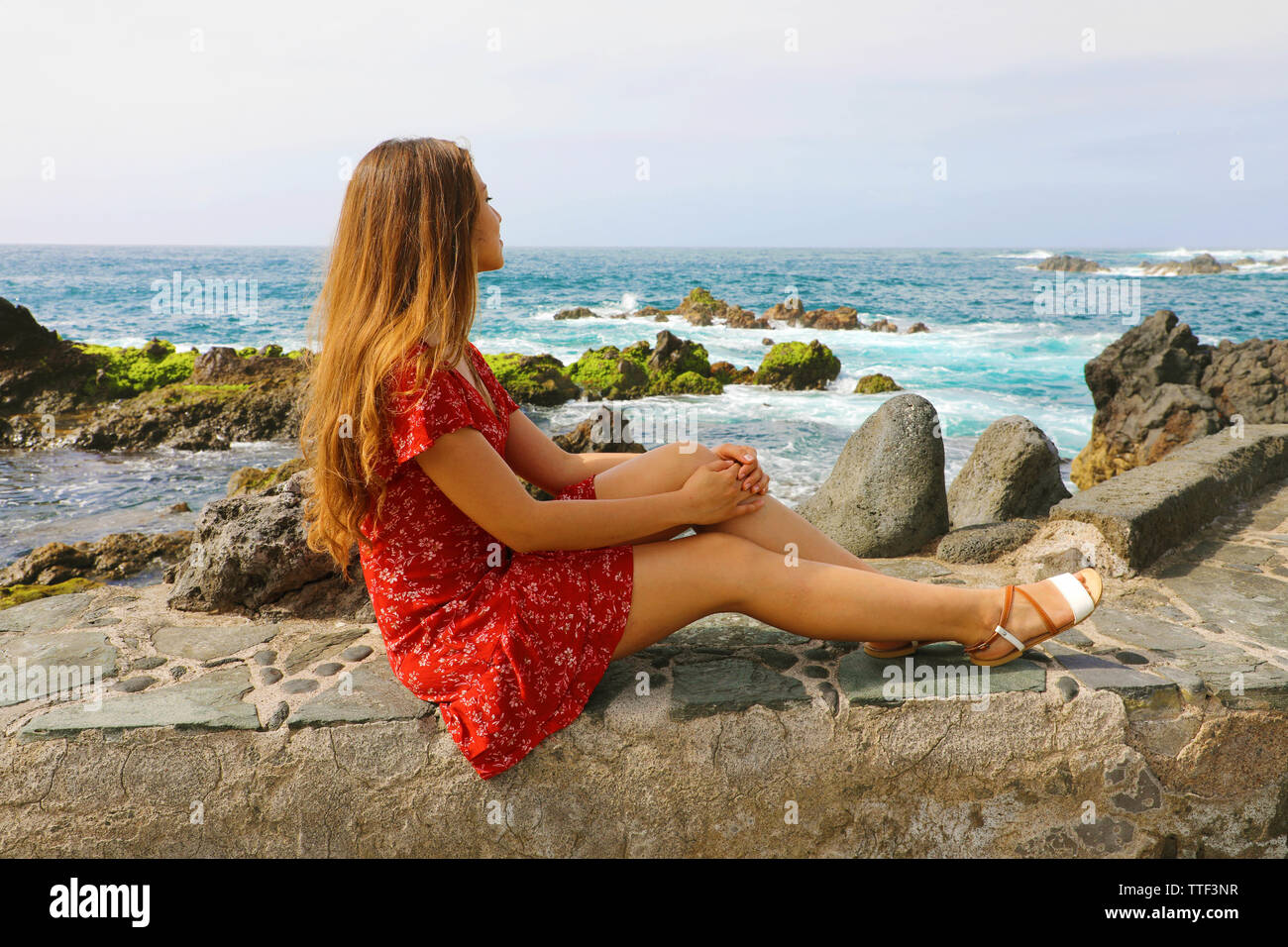 Beautiful young woman sitting on wall looking at stunning seascape in Puerto de la Cruz, Tenerife Stock Photo