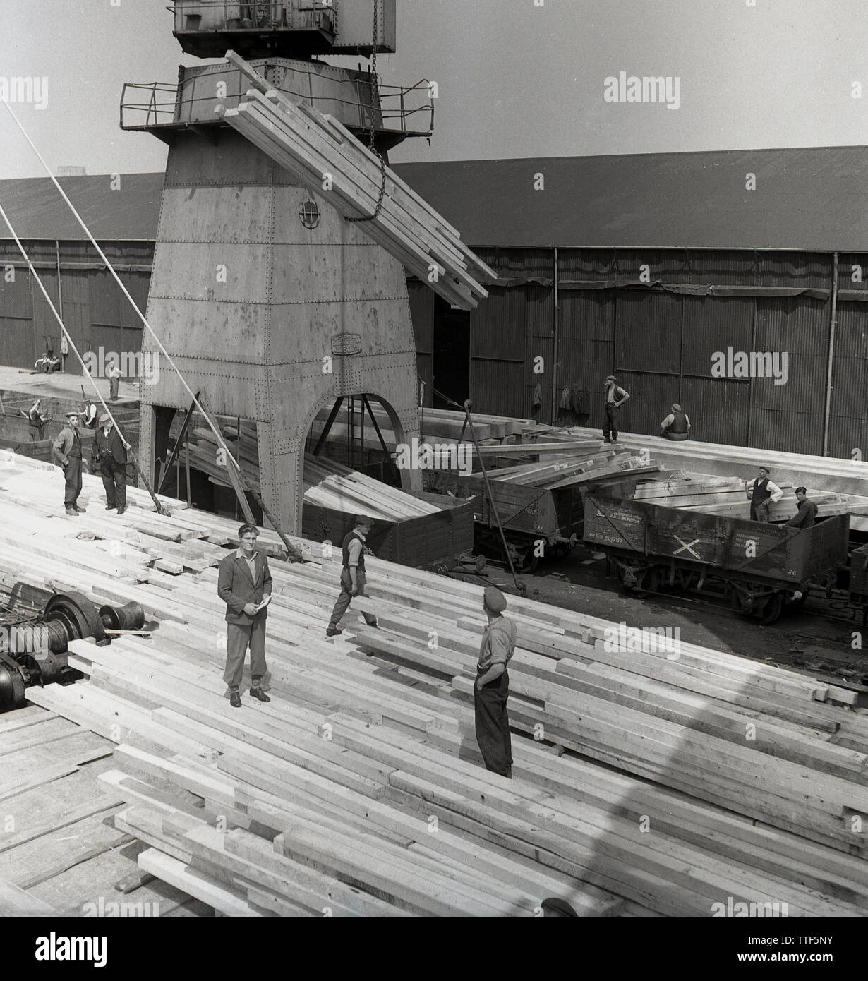 1950s, historical, London docks and a group of dock workers supervising a large cargo of wood being loaded onto GWR freight rail wagons at the docks. Stock Photo