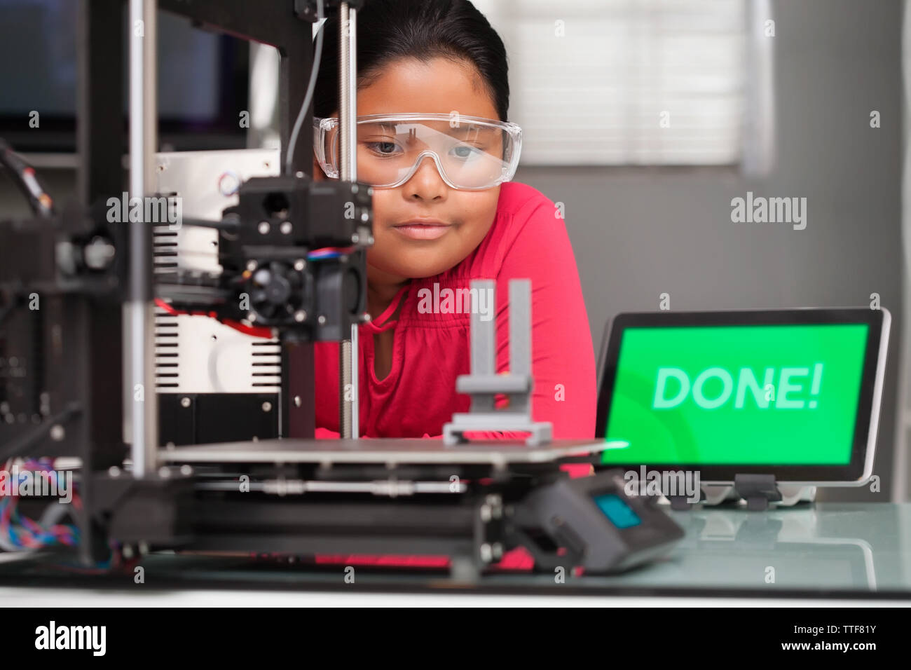 Young girl wearing protective glasses patiently waits for a 3d printer to finish printing her 3d design. Stock Photo