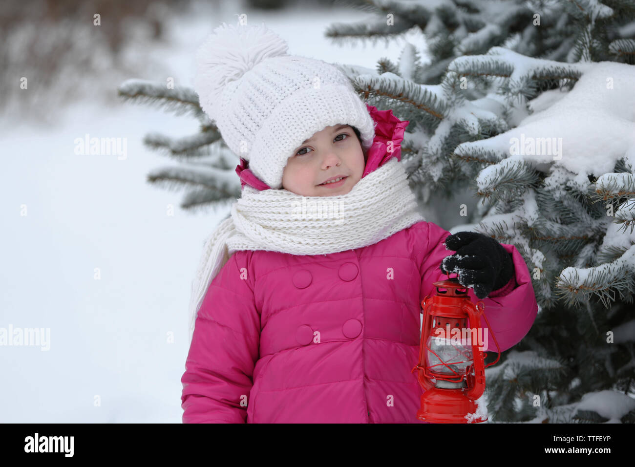 Little girl with winter clothes holding red lantern near fir tree in snowy park outdoor Stock Photo