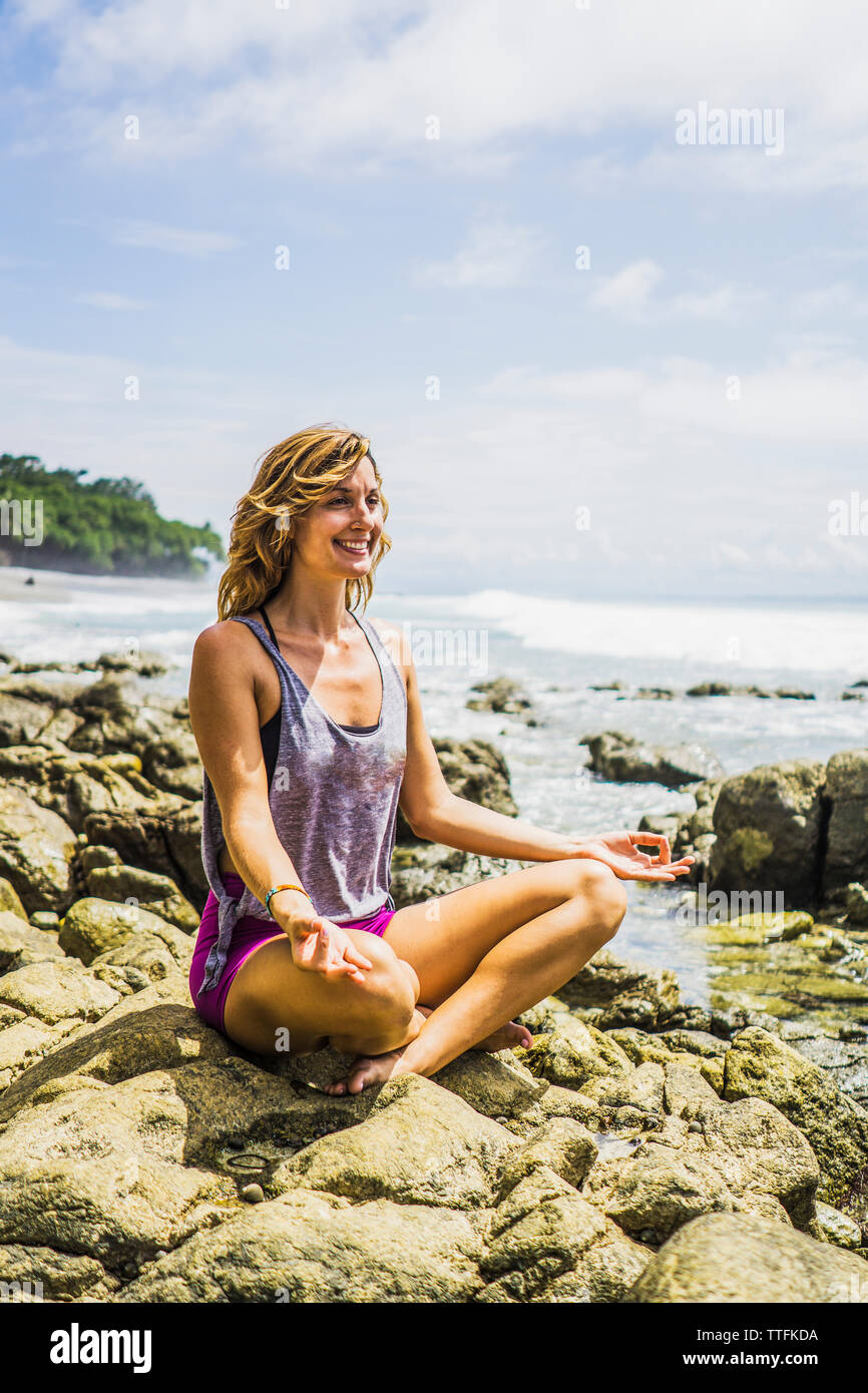 Happy mid adult woman sitting in lotus position on rocks at beach Stock Photo