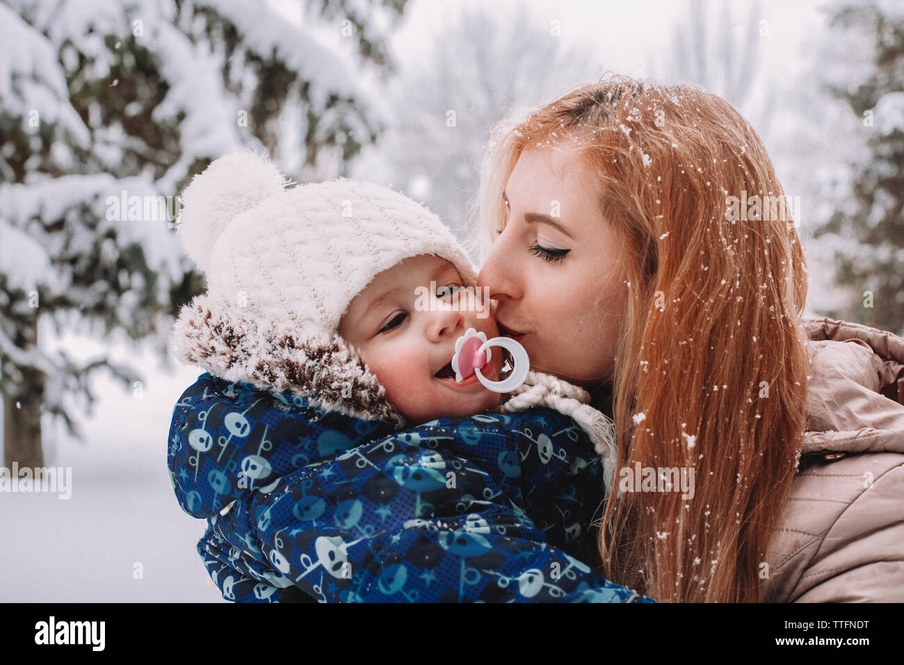 Mother kissing cute son while standing against trees in park during winter Stock Photo