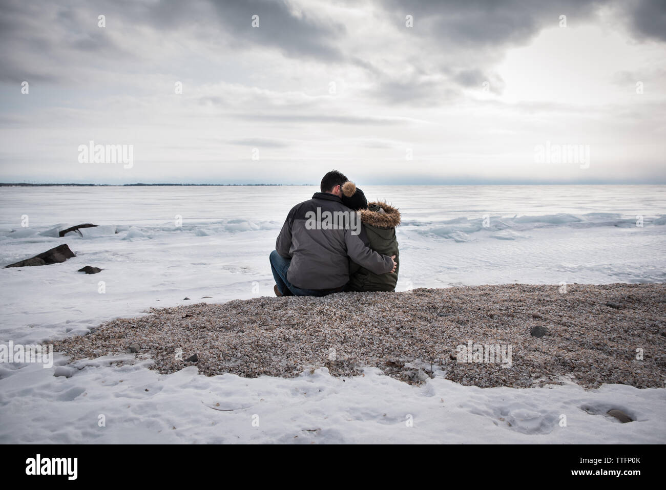 Couple sitting together on icy lake shore on a cloudy winter day. Stock Photo