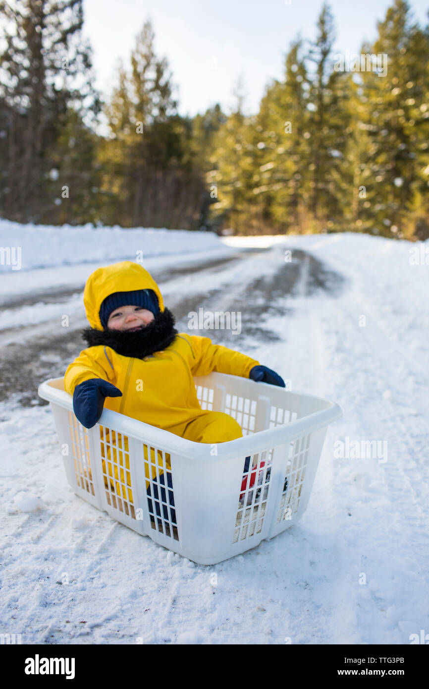 Toddler boy sitting in laundry basket, used as sled in snow Stock Photo