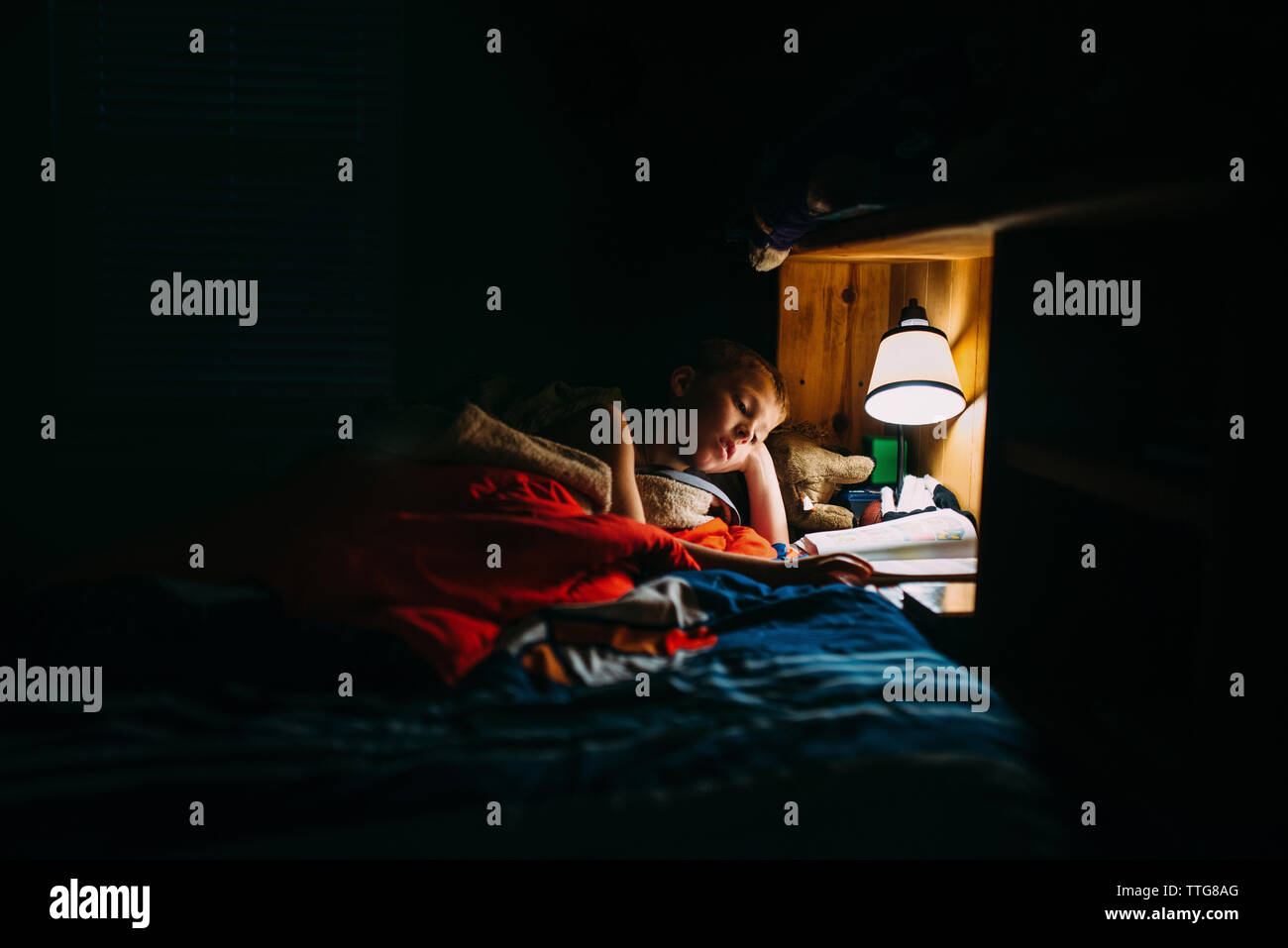 Boy reading book in darkroom under electric lamp at home Stock Photo