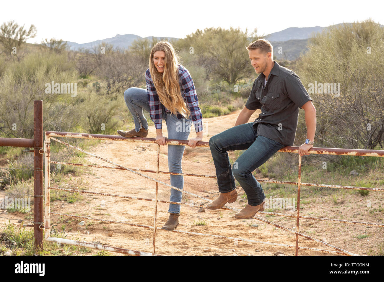 Western wear young couple hopping gate on desert ranch Stock Photo