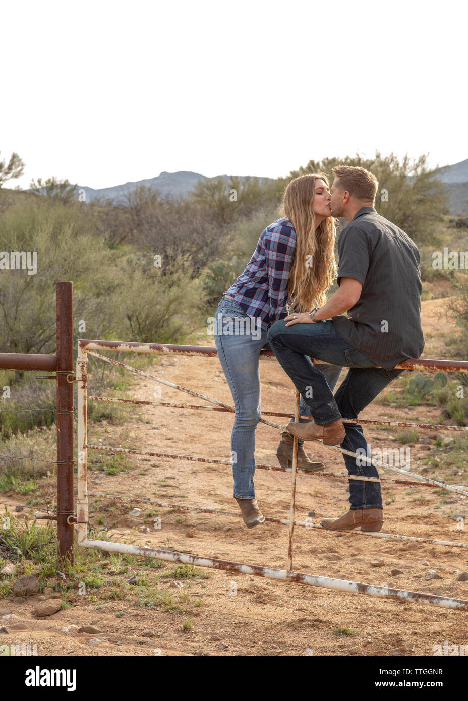 Western wear young couple share a kiss sitting on a ranch gate Stock Photo