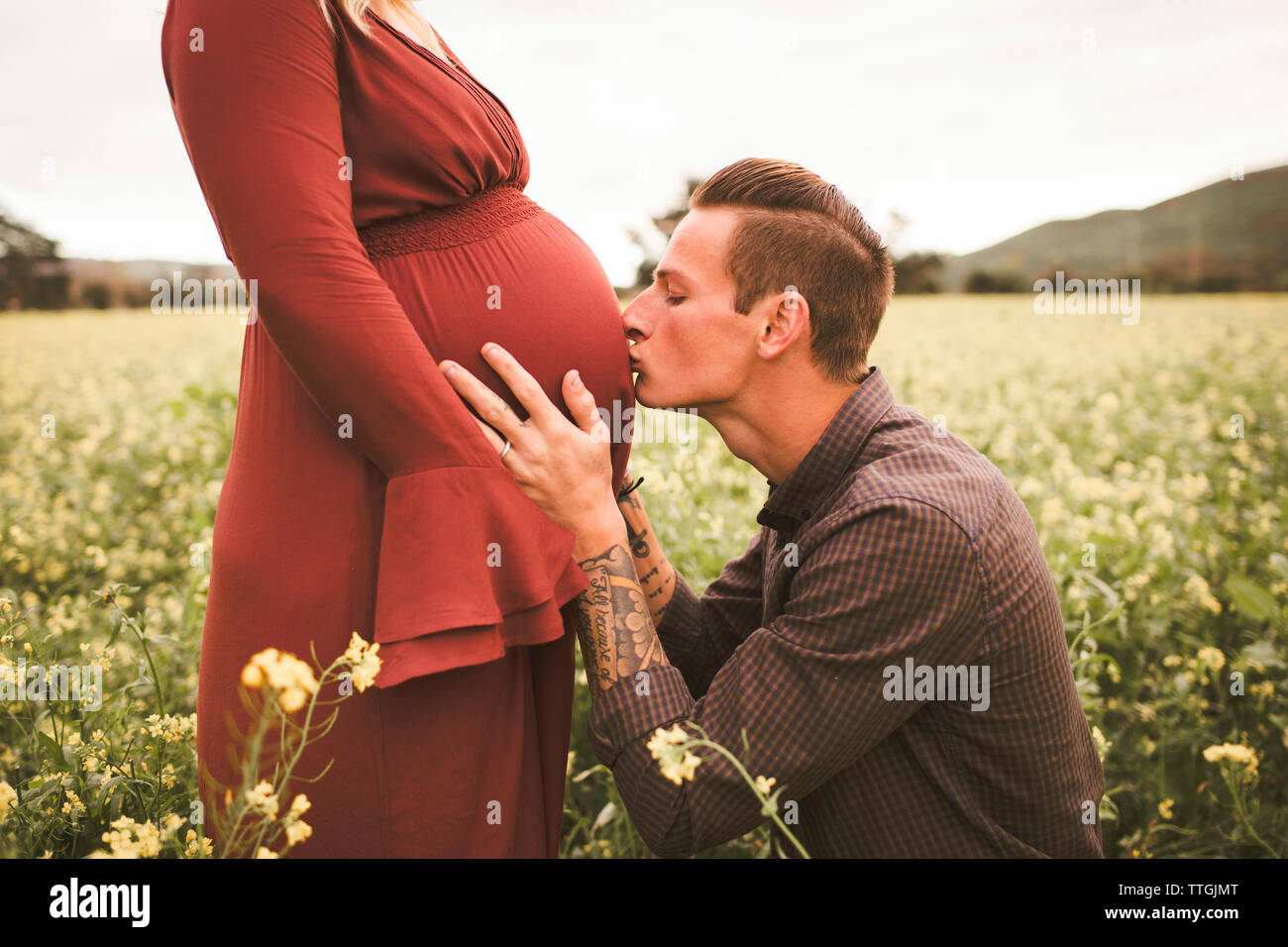 Man kissing pregnant wife's abdomen on field against sky Stock Photo