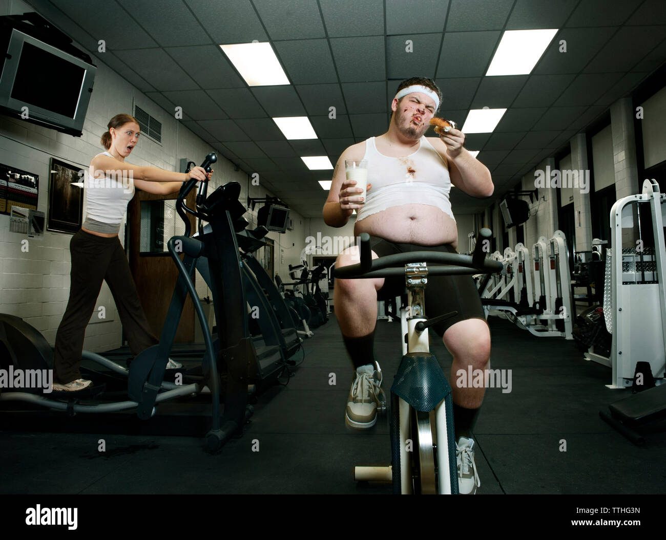Shocked woman looking at fat man eating donut while exercising in gym Stock Photo