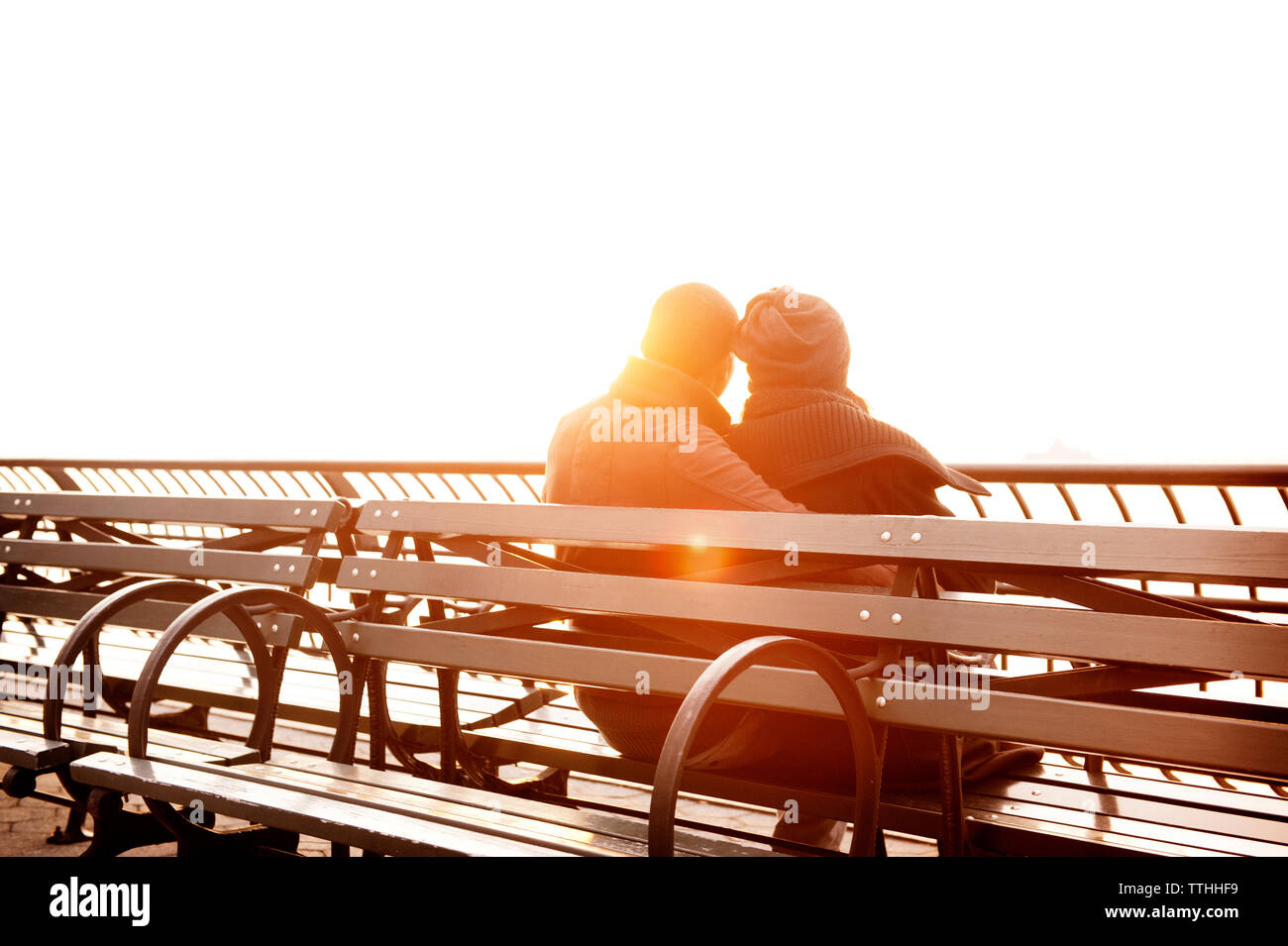 Rear view of couple sitting on bench against sky during sunset Stock Photo