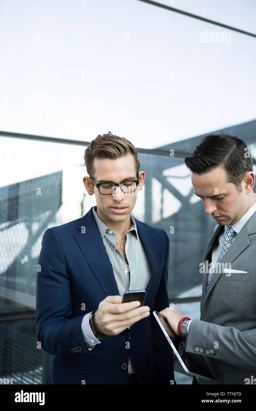 businessman using phone while colleague checking time on watch while standing outdoors Stock Photo