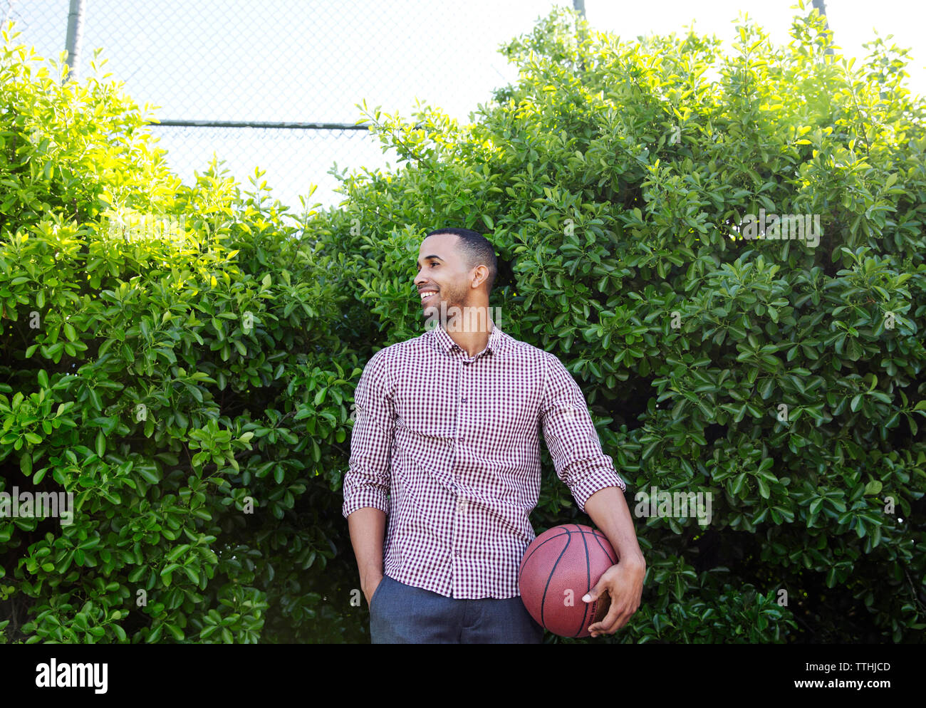 Happy young man holding basketball while standing against plants Stock Photo