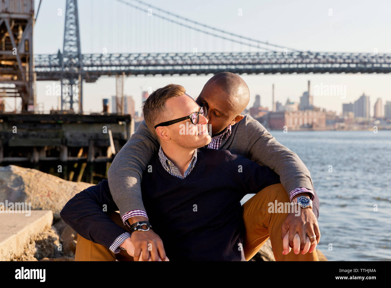 Man kissing boyfriend while sitting against Williamsburg Bridge Stock Photo