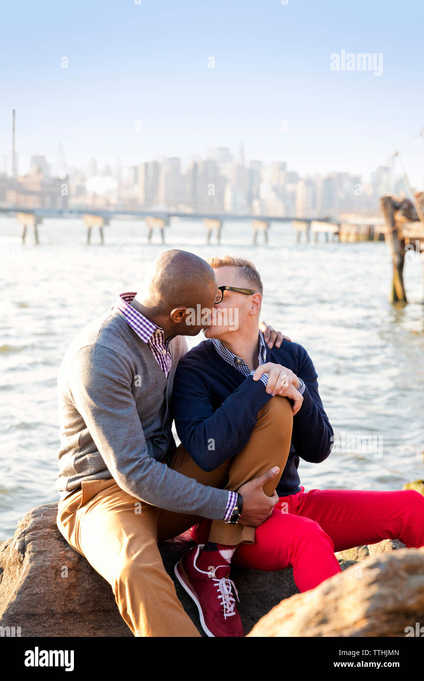 Boyfriends kissing while sitting on rocks against river Stock Photo