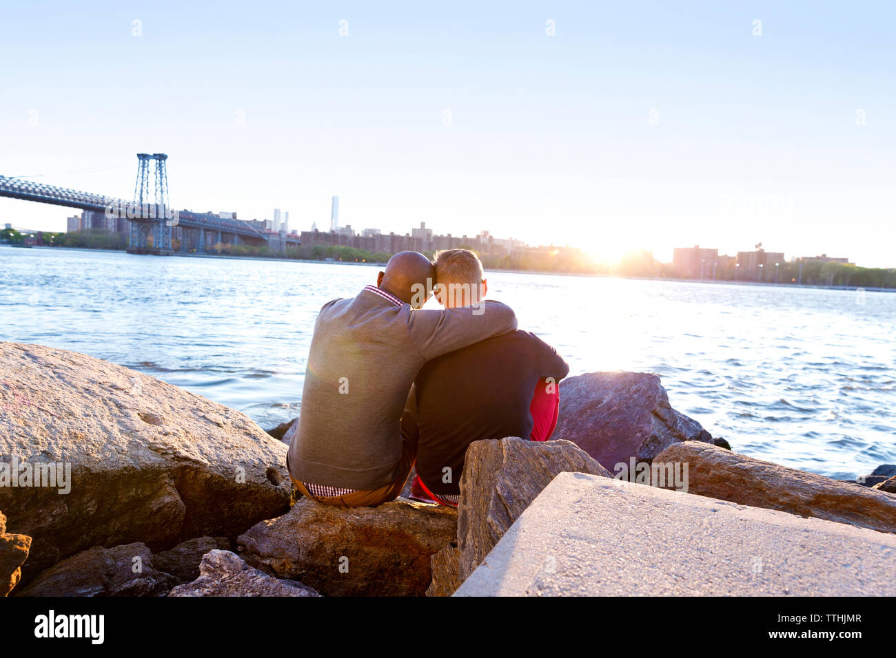 Rear view of man sitting arm around with boyfriend on rocks at East River during sunset Stock Photo