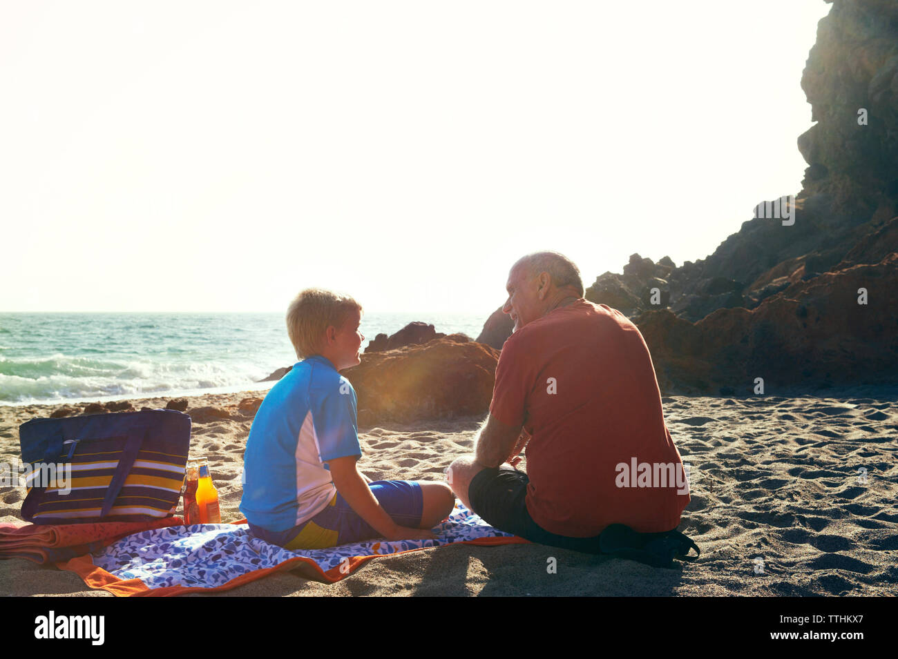 Rear view of senior man talking to grandson while sitting at beach on sunny day Stock Photo