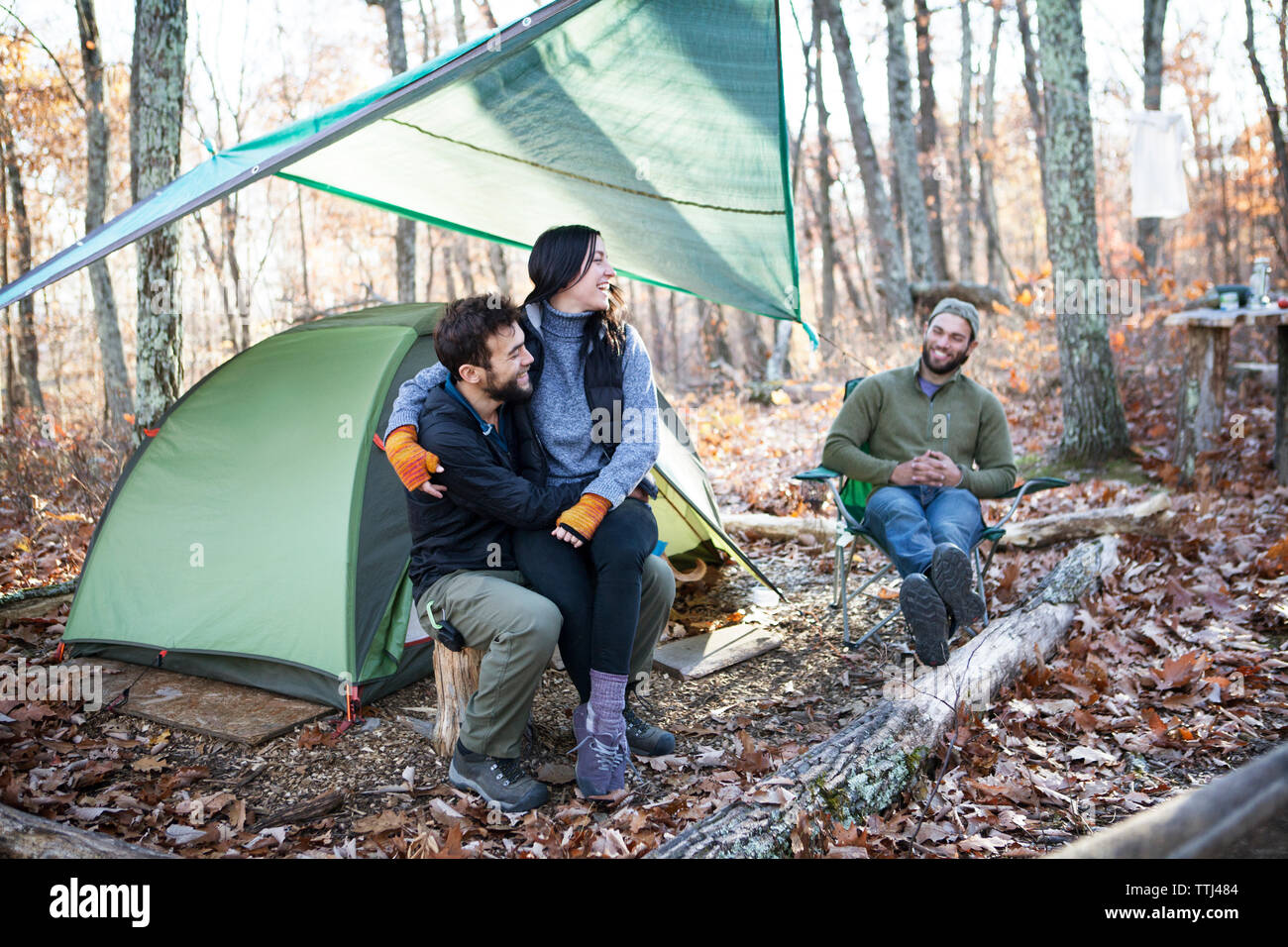 Friend looking at couple while sitting on tree stumps by tent in forest Stock Photo