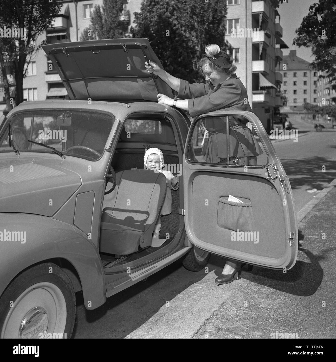 Driving in the 1950s. A young woman in her Fiat convertible car. Note the car door that opens forward and there are no seatbelts. It´s a warm and sunny day and the roof top is removed. Her daughter is seen in the car. Sweden 1951. ref 1711 Stock Photo