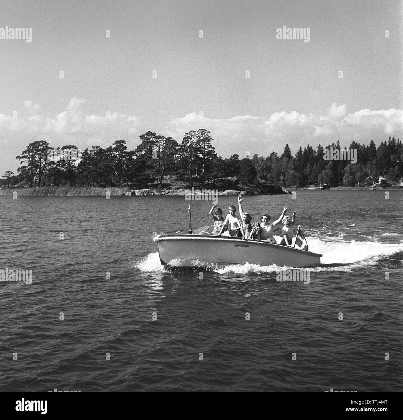 1950s summer. A family passes the photographer in their small motor boat and waves happily.  Sweden 1951  Kristoffersson ref BD57-3. Stock Photo
