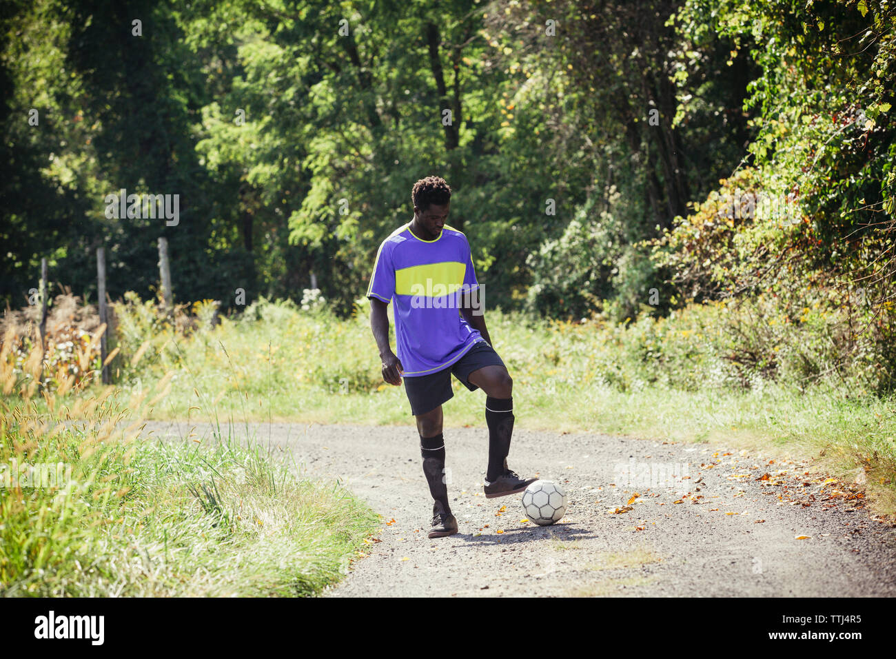 Full length of young man playing with soccer ball on dirt road during sunny day Stock Photo