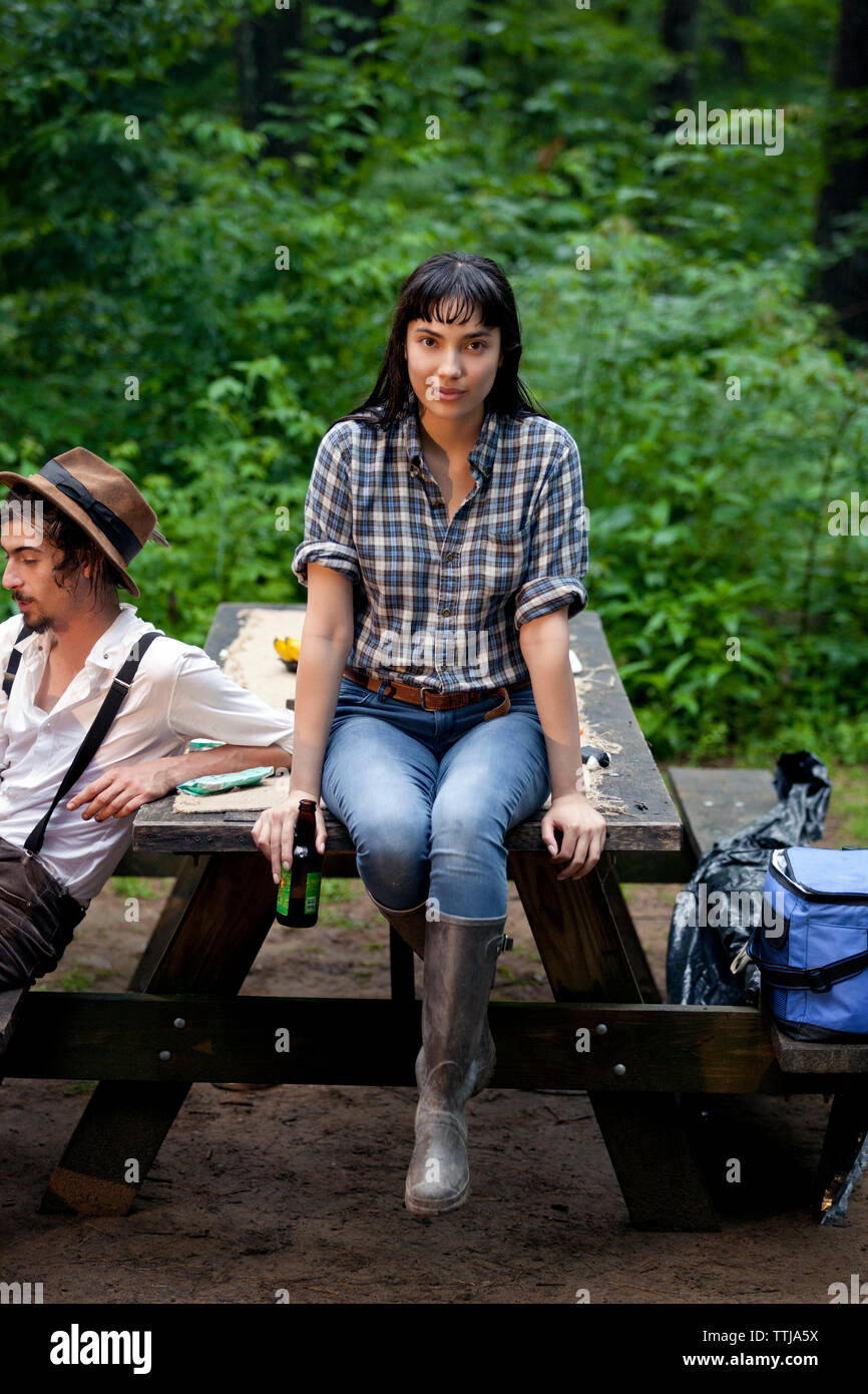 Portrait of woman holding beer bottle while sitting with friend on picnic table in forest Stock Photo