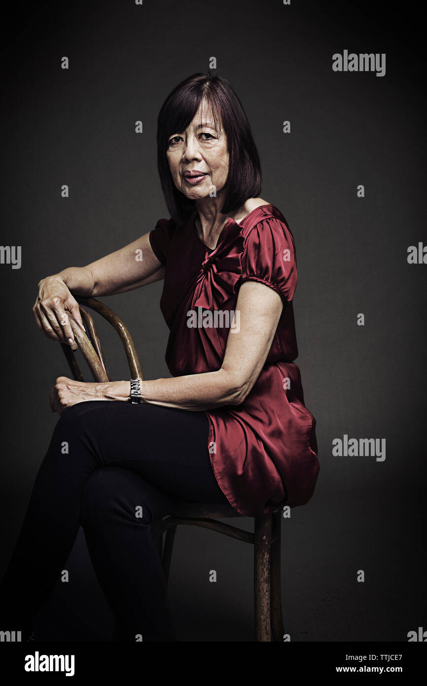 Portrait of senior female sitting on chair against black background ...