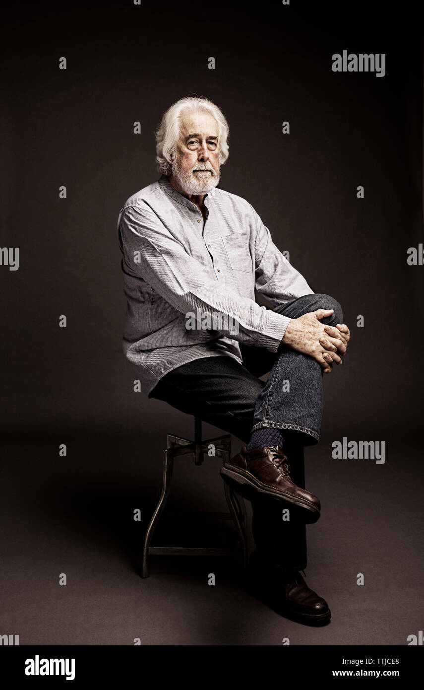 Portrait of senior man with white hair sitting on stool against black background Stock Photo