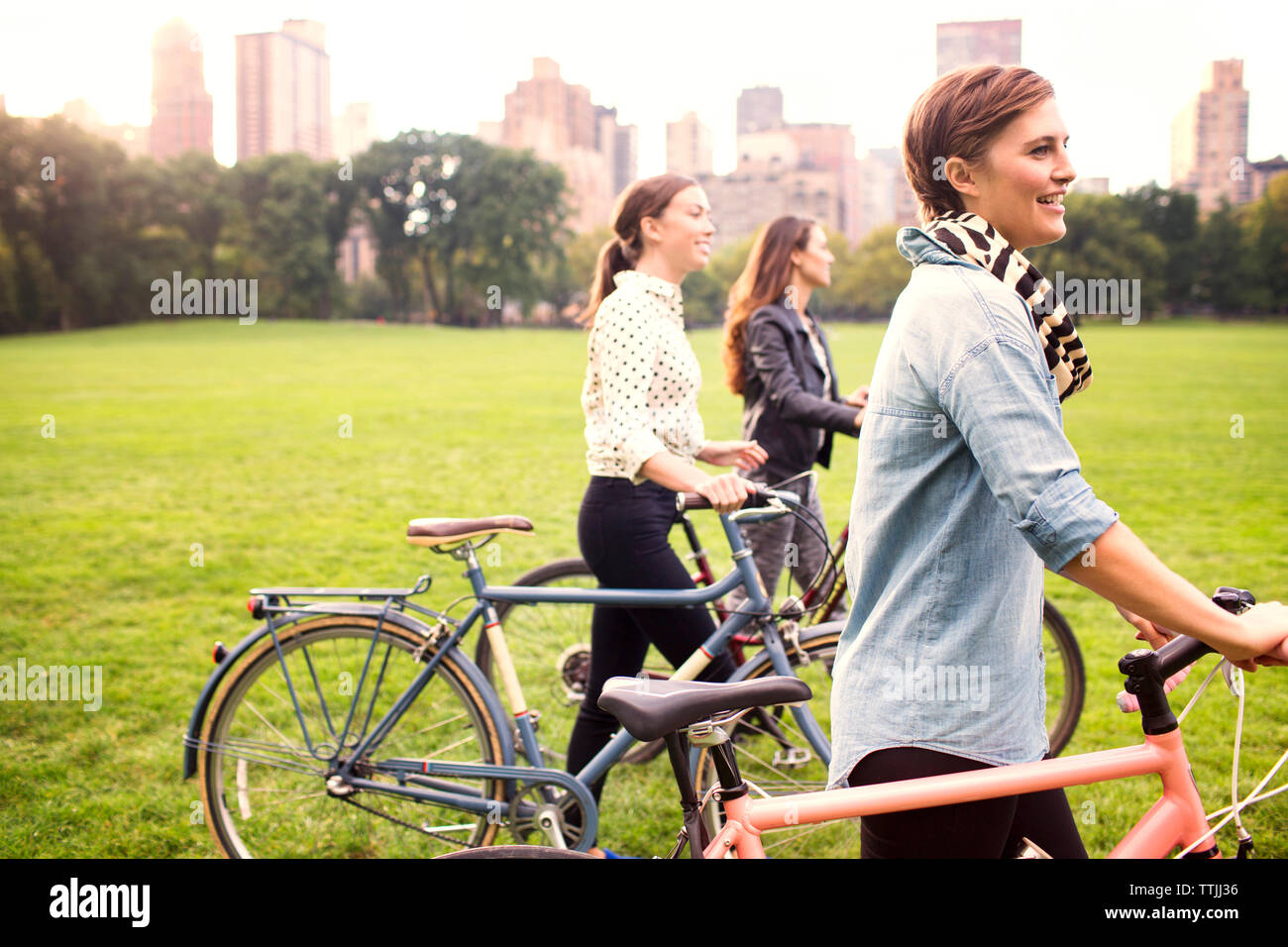 Women with bicycles walking on field in park against city Stock Photo
