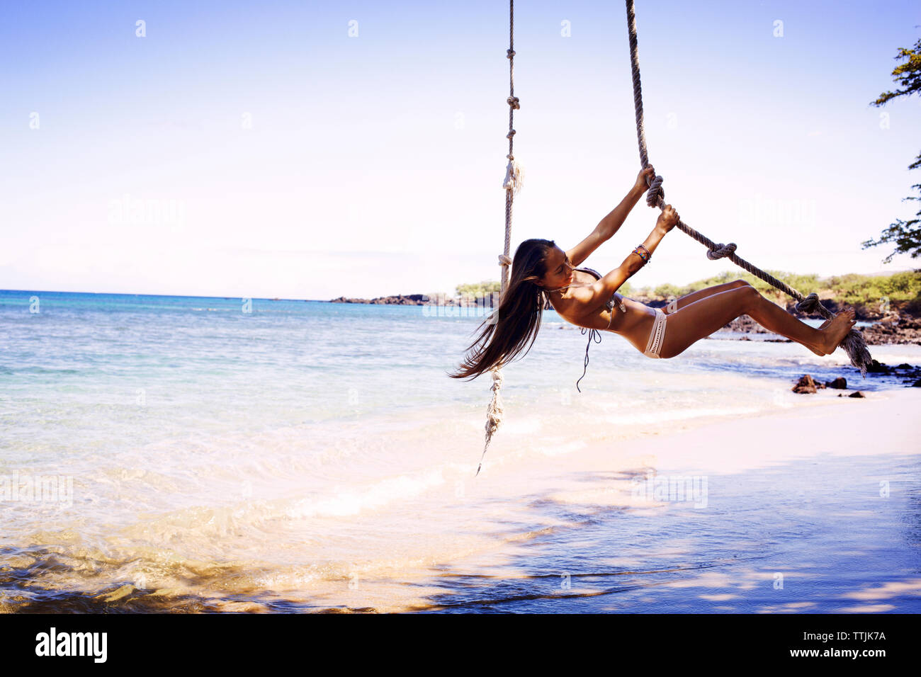 Woman in bikini swinging on rope at beach against clear sky Stock Photo