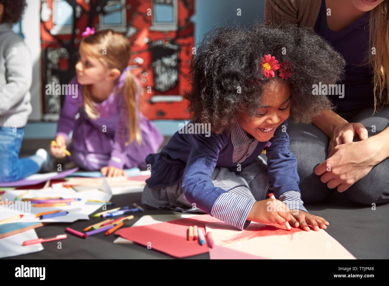 Cheerful girl drawing on paper while sitting by teacher at preschool Stock Photo