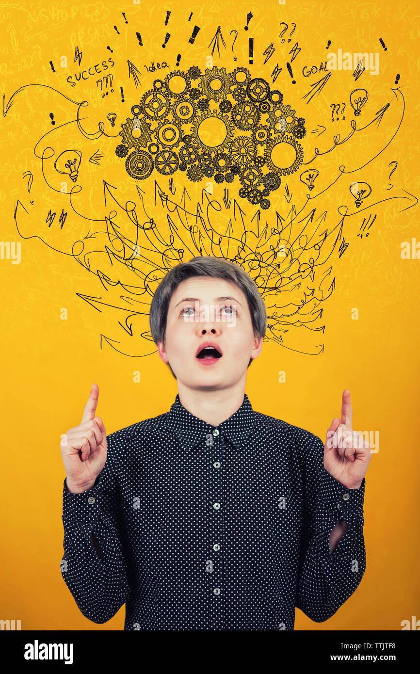 Young woman points forefingers up, looking astonished and curious with mouth open at cogwheel brain above head. Hard thinking, arrows and curves mess Stock Photo