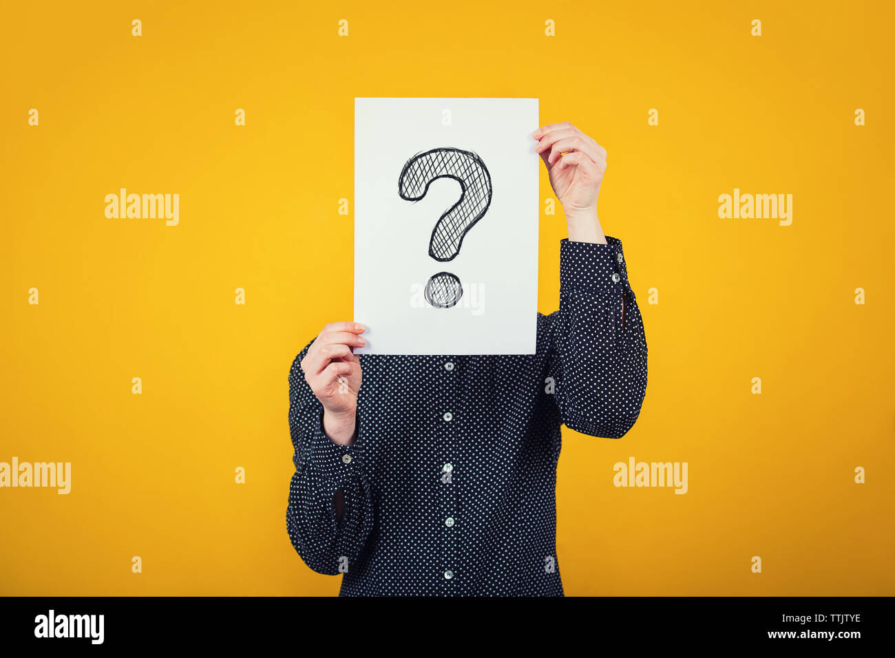 Businesswoman covering face using a white paper sheet with drawn question mark, like a mask, for hiding her identity. Isolated on yellow wall backgrou Stock Photo
