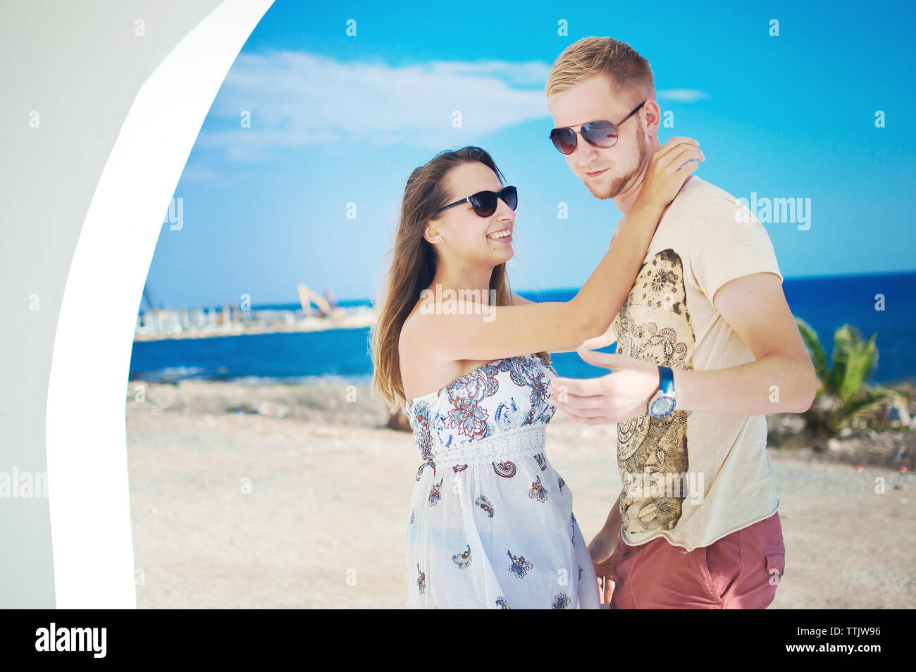 Image of young man in sunglasses holding and hugging caucassian girl in long white dress with brown hair, smiling, looking each other in the eye. Warm Stock Photo