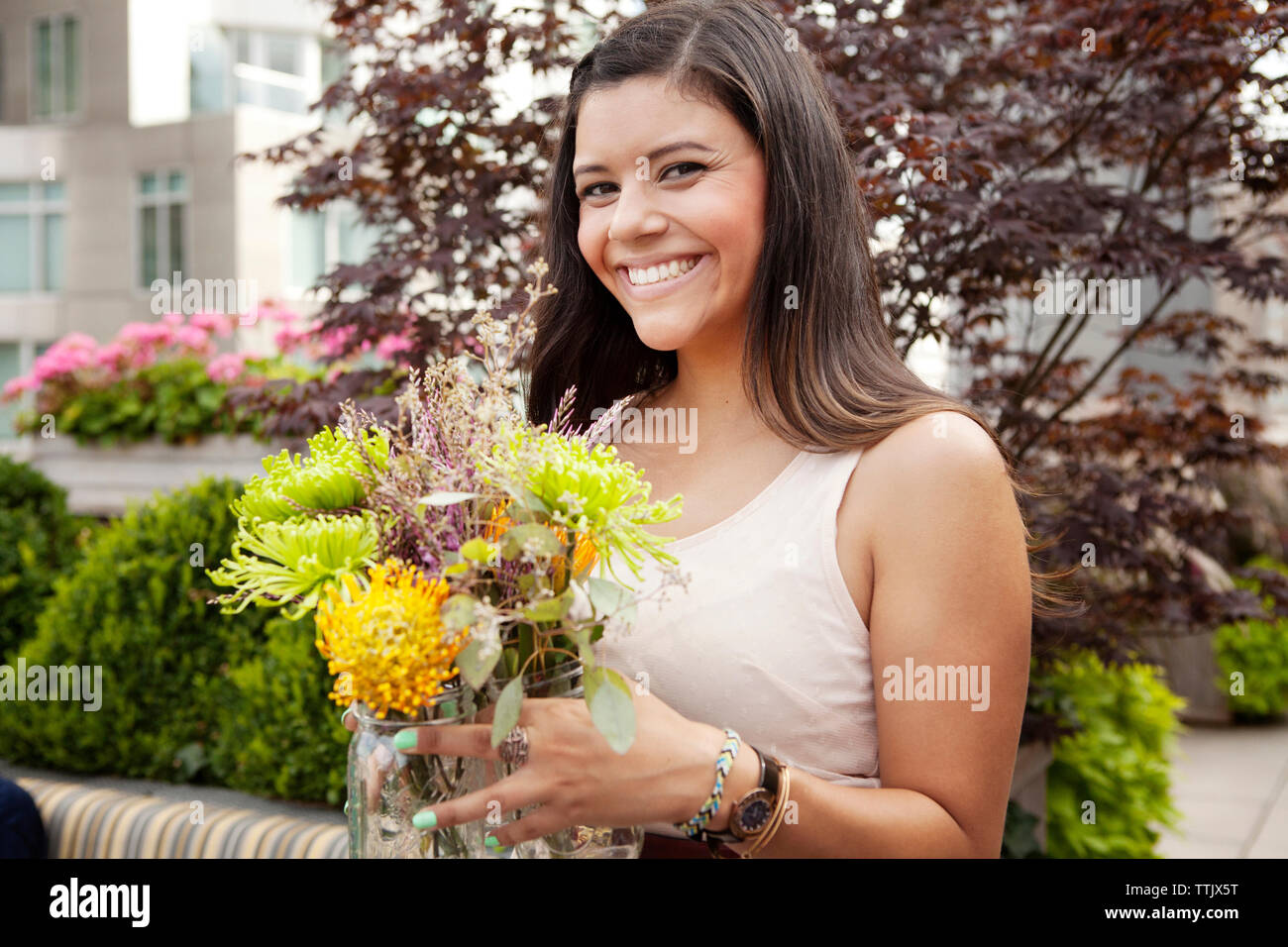 Smiling woman holding flowers jar while standing against plants Stock Photo