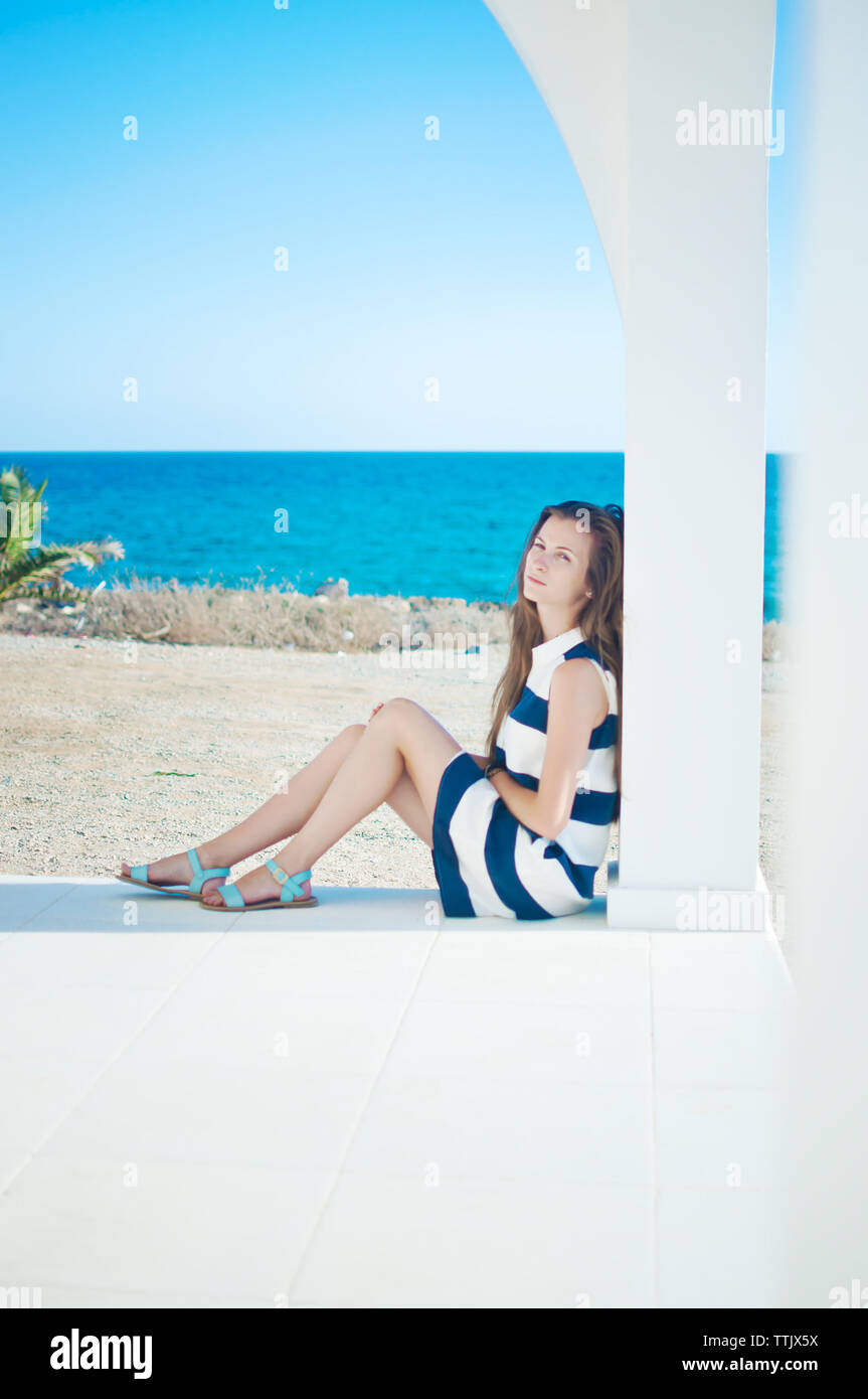Closeup of caucassian girl in striped dress and sandals with long brown hair sitting, leaning against column, looking in the camera. Warm summer day. Stock Photo